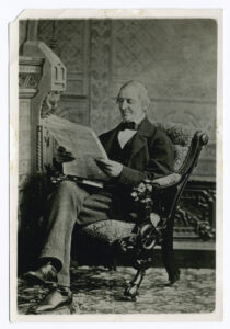 Black and white photo of Emerson reading a newspaper. He wears a coat and bowtie and sits in an ornate chair indoors.